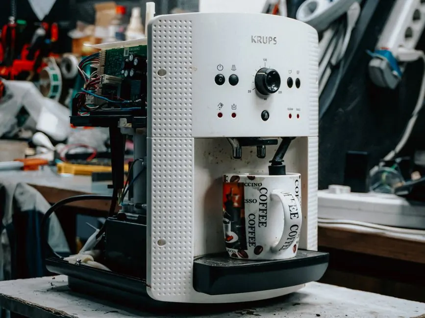 A coffee machine undergoing repair in a cluttered workshop, with parts exposed and a coffee mug.