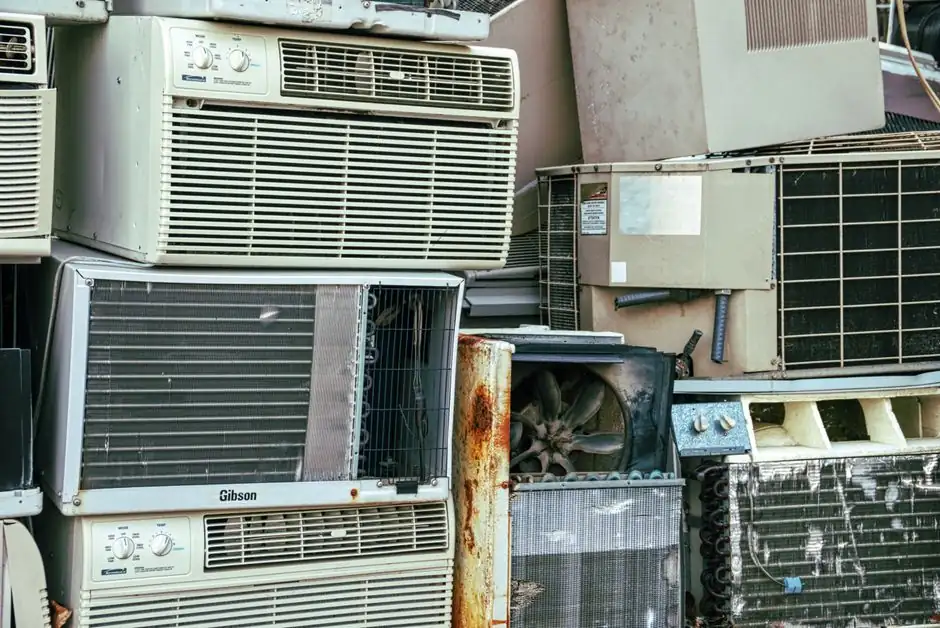 A collection of broken and rusted air conditioners piled in an outdoor junkyard.